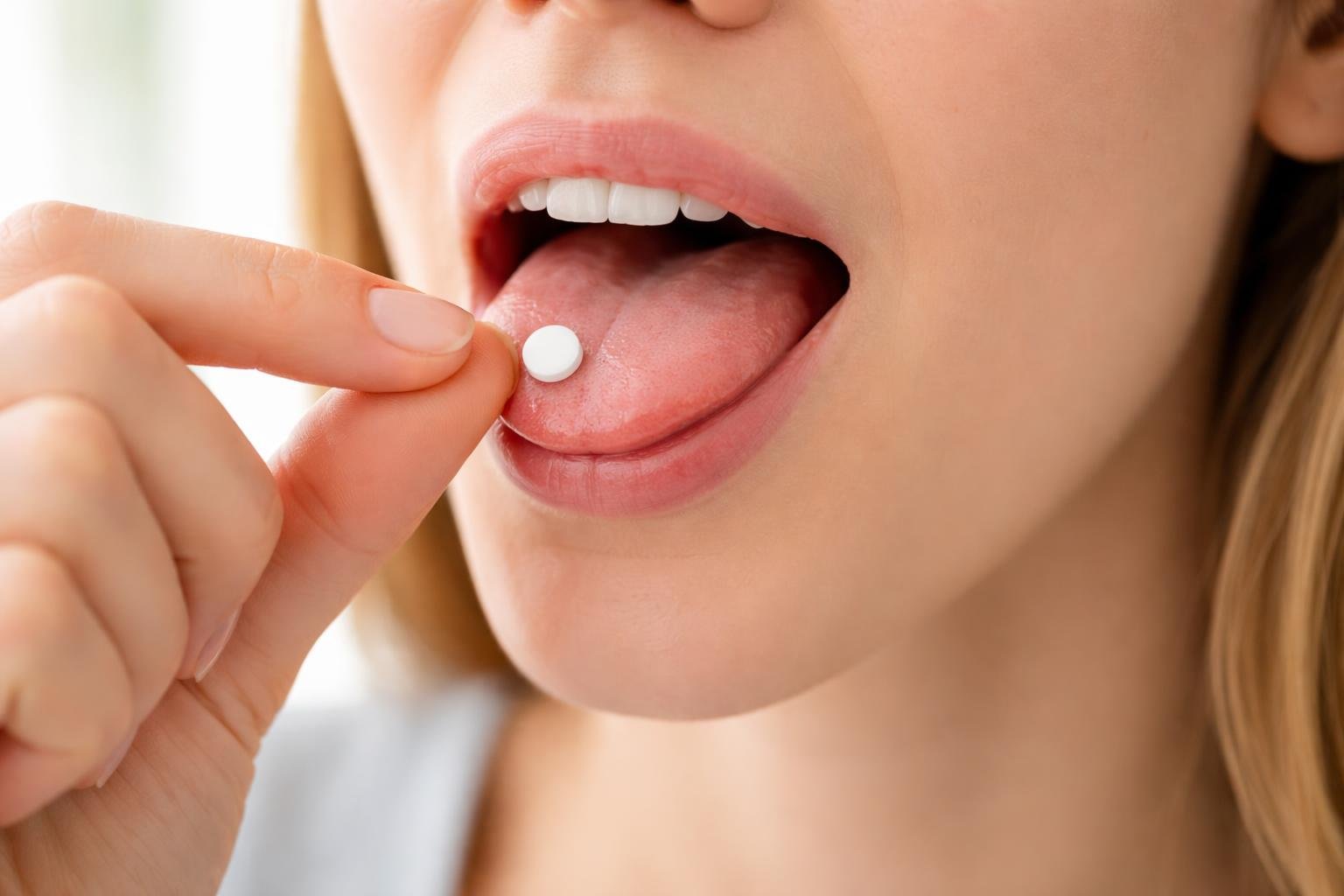 Pessoa segurando um comprimido sublingual próximo à boca aberta, com a língua levemente levantada para colocar o remédio embaixo.
